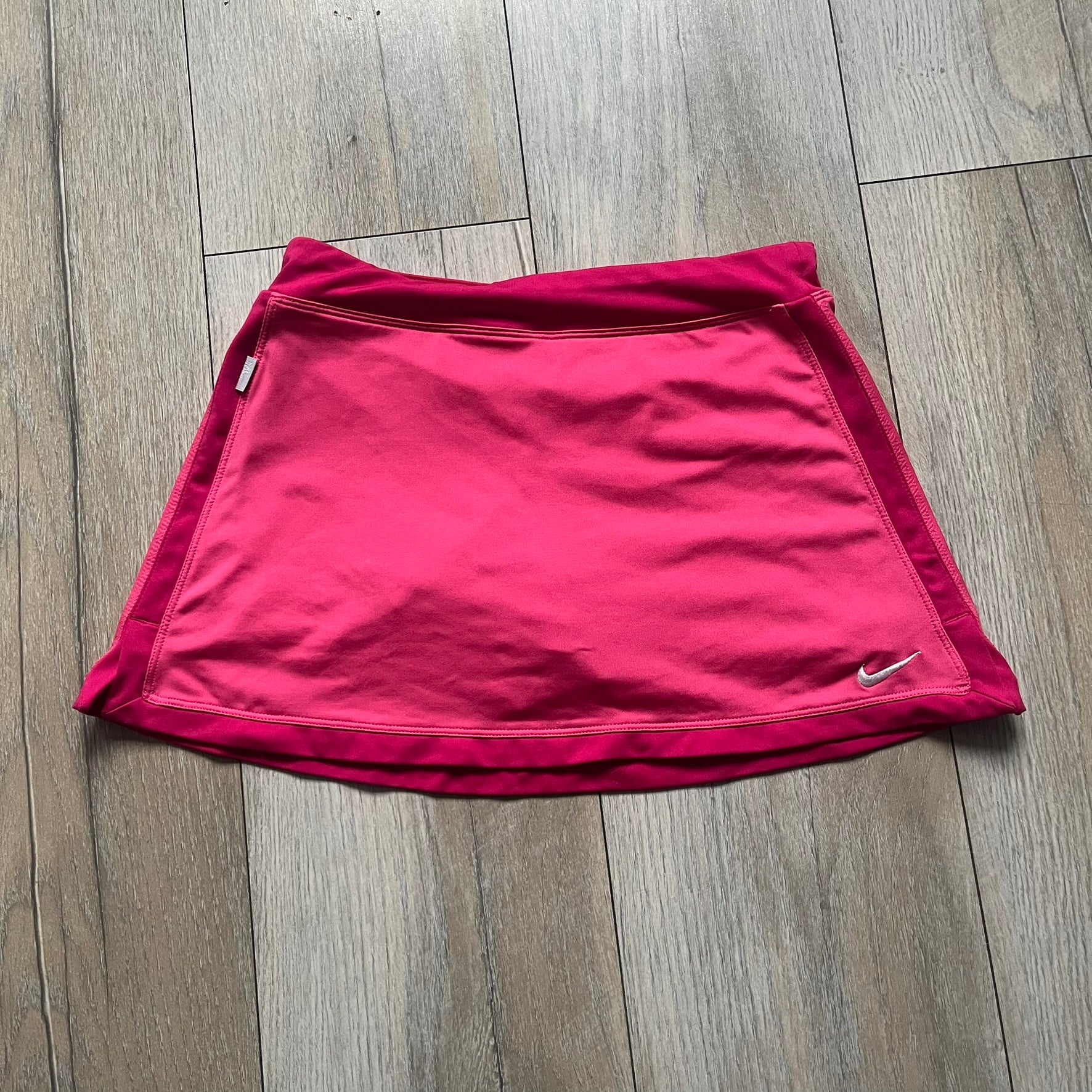 Nike Dri-FIT Pink Tennis Skirt is a women’s tennis skort with built-in shorts for coverage and support. The skirt is finished in dark pink with darker side panels and waistband, featuring a white embroidered Nike Swoosh at the lower hem. A small Nike Court logo on the side highlights its connection to Nike’s performance tennis line.