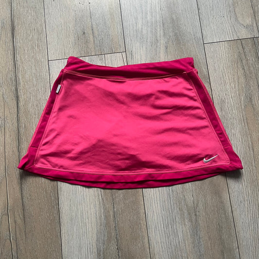 Nike Dri-FIT Pink Tennis Skirt is a women’s tennis skort with built-in shorts for coverage and support. The skirt is finished in dark pink with darker side panels and waistband, featuring a white embroidered Nike Swoosh at the lower hem. A small Nike Court logo on the side highlights its connection to Nike’s performance tennis line.