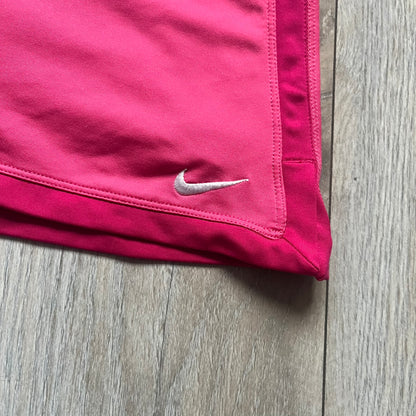 Nike Dri-FIT Pink Tennis Skirt is a women’s tennis skort with built-in shorts for coverage and support. The skirt is finished in dark pink with darker side panels and waistband, featuring a white embroidered Nike Swoosh at the lower hem. A small Nike Court logo on the side highlights its connection to Nike’s performance tennis line.