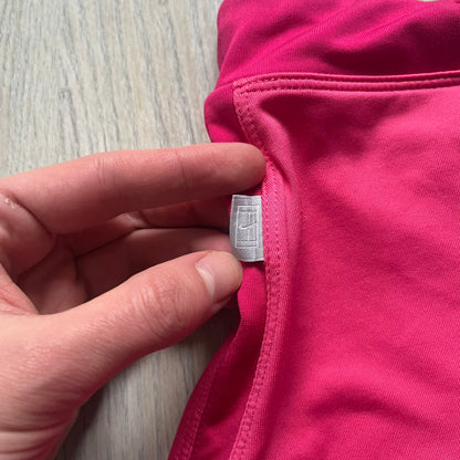 Nike Dri-FIT Pink Tennis Skirt is a women’s tennis skort with built-in shorts for coverage and support. The skirt is finished in dark pink with darker side panels and waistband, featuring a white embroidered Nike Swoosh at the lower hem. A small Nike Court logo on the side highlights its connection to Nike’s performance tennis line.