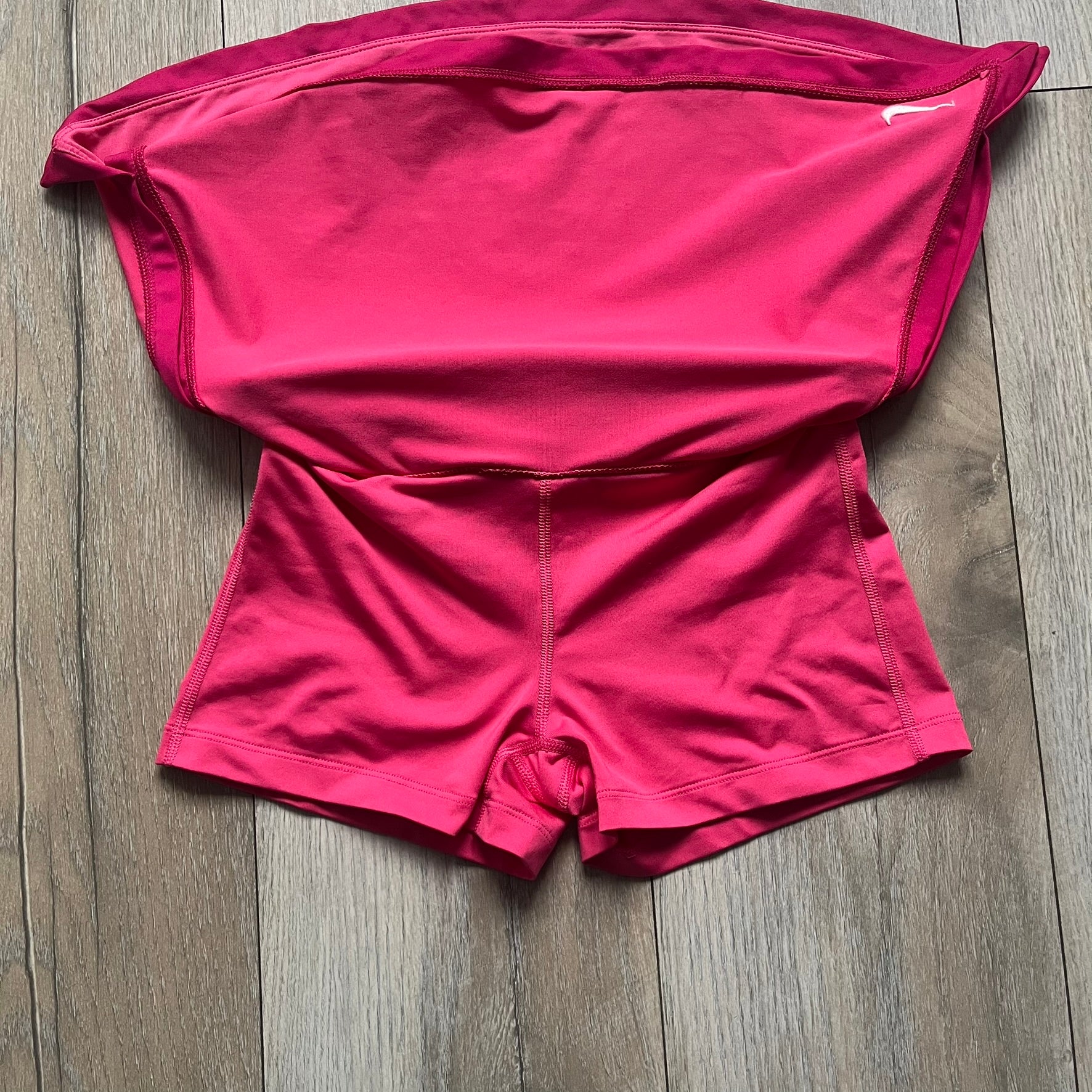 Nike Dri-FIT Pink Tennis Skirt is a women’s tennis skort with built-in shorts for coverage and support. The skirt is finished in dark pink with darker side panels and waistband, featuring a white embroidered Nike Swoosh at the lower hem. A small Nike Court logo on the side highlights its connection to Nike’s performance tennis line.