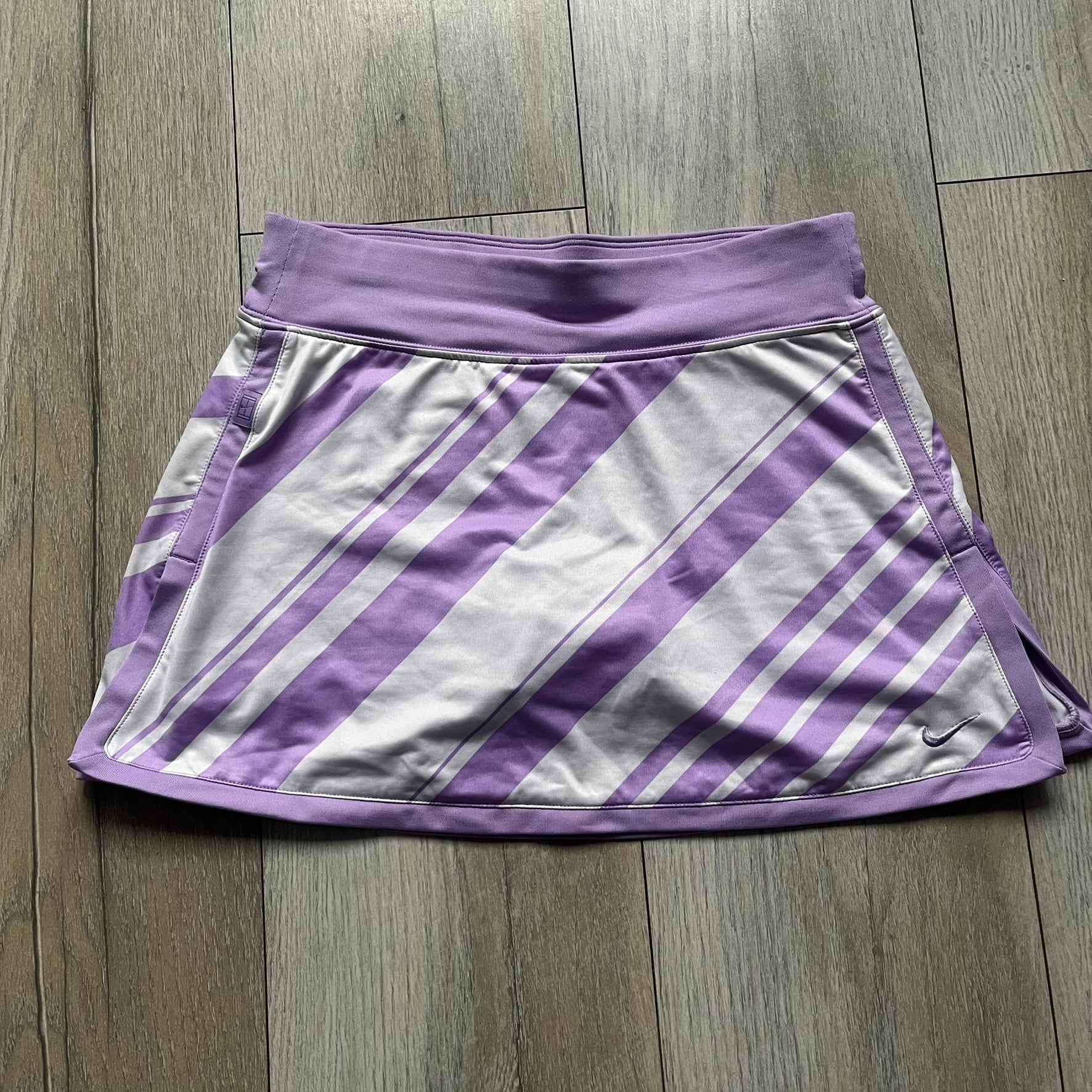 Nike Dri-FIT Striped Lavender Tennis Skirt is a women’s tennis skort with integrated inner shorts for coverage and support. The skirt features diagonal white and lavender stripes on the front and back, a small Nike Court logo on the side, and an embroidered lavender Swoosh at the lower front. The lavender waistband and side cutouts provide comfort and mobility during play.