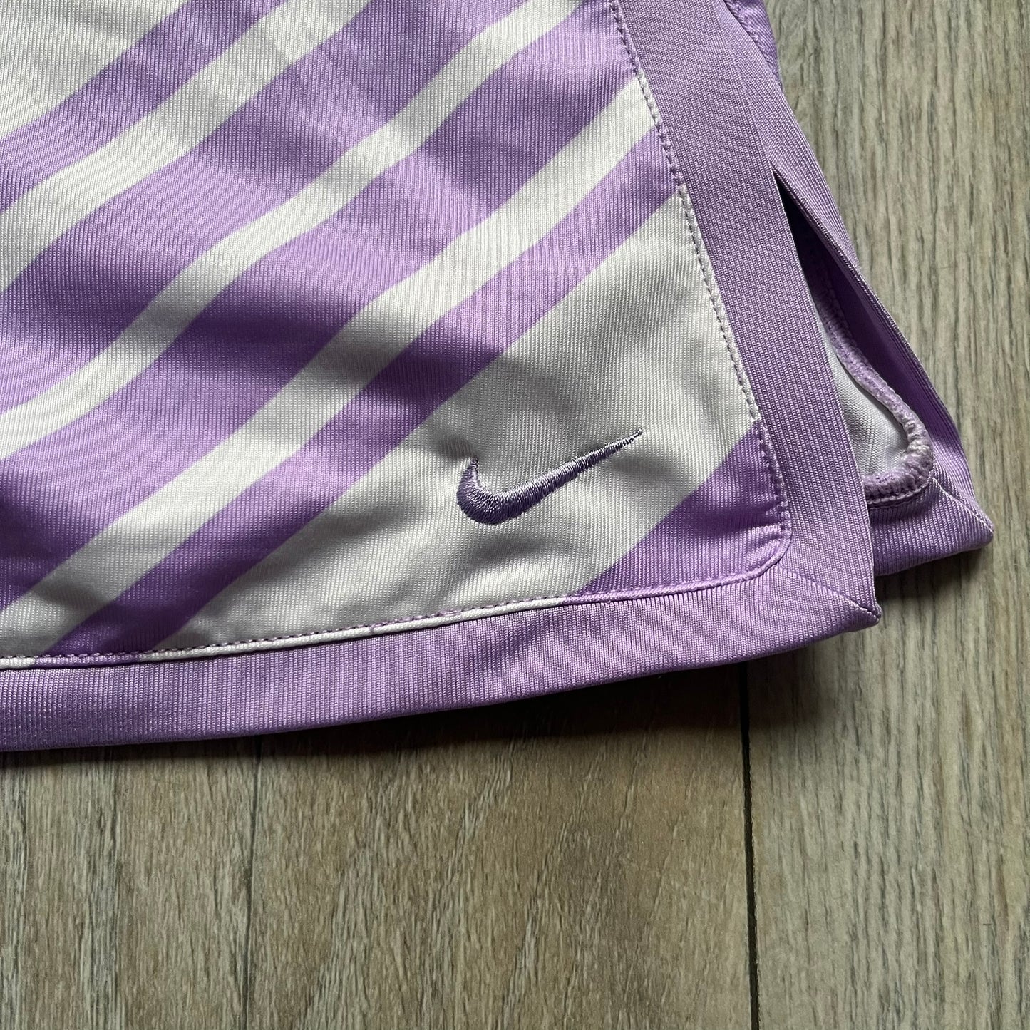 Nike Dri-FIT Striped Lavender Tennis Skirt is a women’s tennis skort with integrated inner shorts for coverage and support. The skirt features diagonal white and lavender stripes on the front and back, a small Nike Court logo on the side, and an embroidered lavender Swoosh at the lower front. The lavender waistband and side cutouts provide comfort and mobility during play.