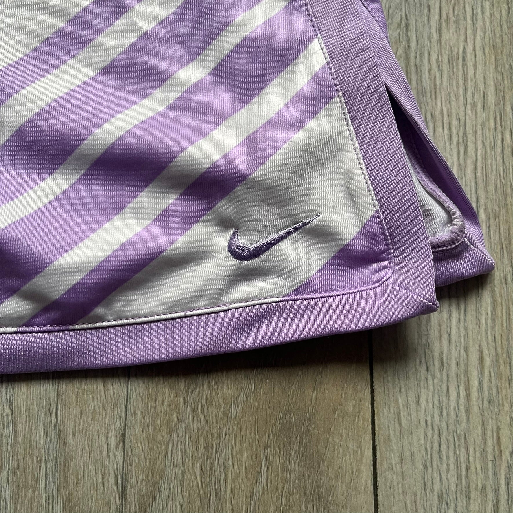 Nike Dri-FIT Striped Lavender Tennis Skirt is a women’s tennis skort with integrated inner shorts for coverage and support. The skirt features diagonal white and lavender stripes on the front and back, a small Nike Court logo on the side, and an embroidered lavender Swoosh at the lower front. The lavender waistband and side cutouts provide comfort and mobility during play.