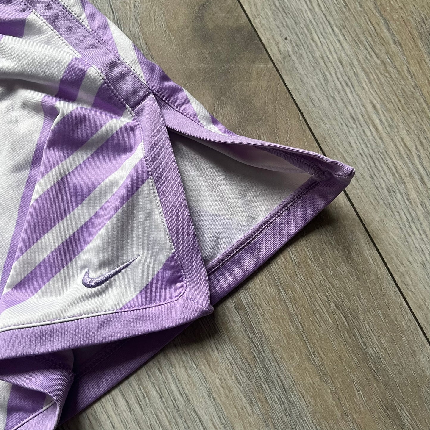 Nike Dri-FIT Striped Lavender Tennis Skirt is a women’s tennis skort with integrated inner shorts for coverage and support. The skirt features diagonal white and lavender stripes on the front and back, a small Nike Court logo on the side, and an embroidered lavender Swoosh at the lower front. The lavender waistband and side cutouts provide comfort and mobility during play.