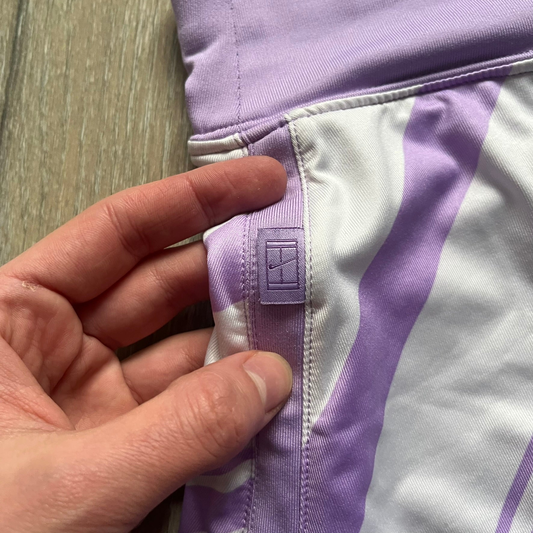 Nike Dri-FIT Striped Lavender Tennis Skirt is a women’s tennis skort with integrated inner shorts for coverage and support. The skirt features diagonal white and lavender stripes on the front and back, a small Nike Court logo on the side, and an embroidered lavender Swoosh at the lower front. The lavender waistband and side cutouts provide comfort and mobility during play.