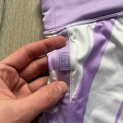 Nike Dri-FIT Striped Lavender Tennis Skirt is a women’s tennis skort with integrated inner shorts for coverage and support. The skirt features diagonal white and lavender stripes on the front and back, a small Nike Court logo on the side, and an embroidered lavender Swoosh at the lower front. The lavender waistband and side cutouts provide comfort and mobility during play.