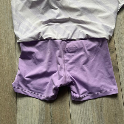Nike Dri-FIT Striped Lavender Tennis Skirt is a women’s tennis skort with integrated inner shorts for coverage and support. The skirt features diagonal white and lavender stripes on the front and back, a small Nike Court logo on the side, and an embroidered lavender Swoosh at the lower front. The lavender waistband and side cutouts provide comfort and mobility during play.