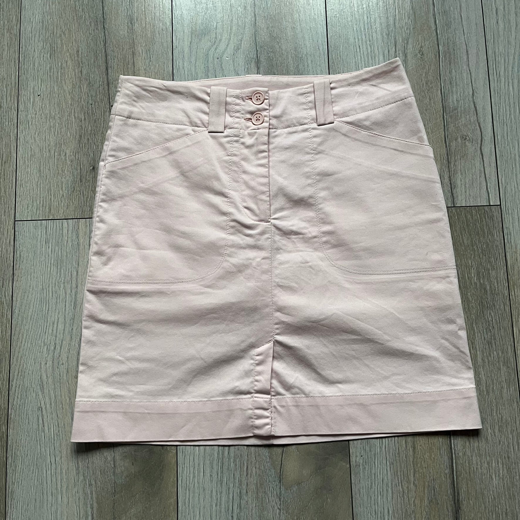 Nike Fit-Dry Light Pink Golf Skirt is a women’s golf skort with integrated inner shorts. The light pink skirt features belt loops at the waistband, front and back pockets, side cutouts for flexibility, and a button closure. A small embroidered Nike logo is placed at the back, maintaining a minimal design.