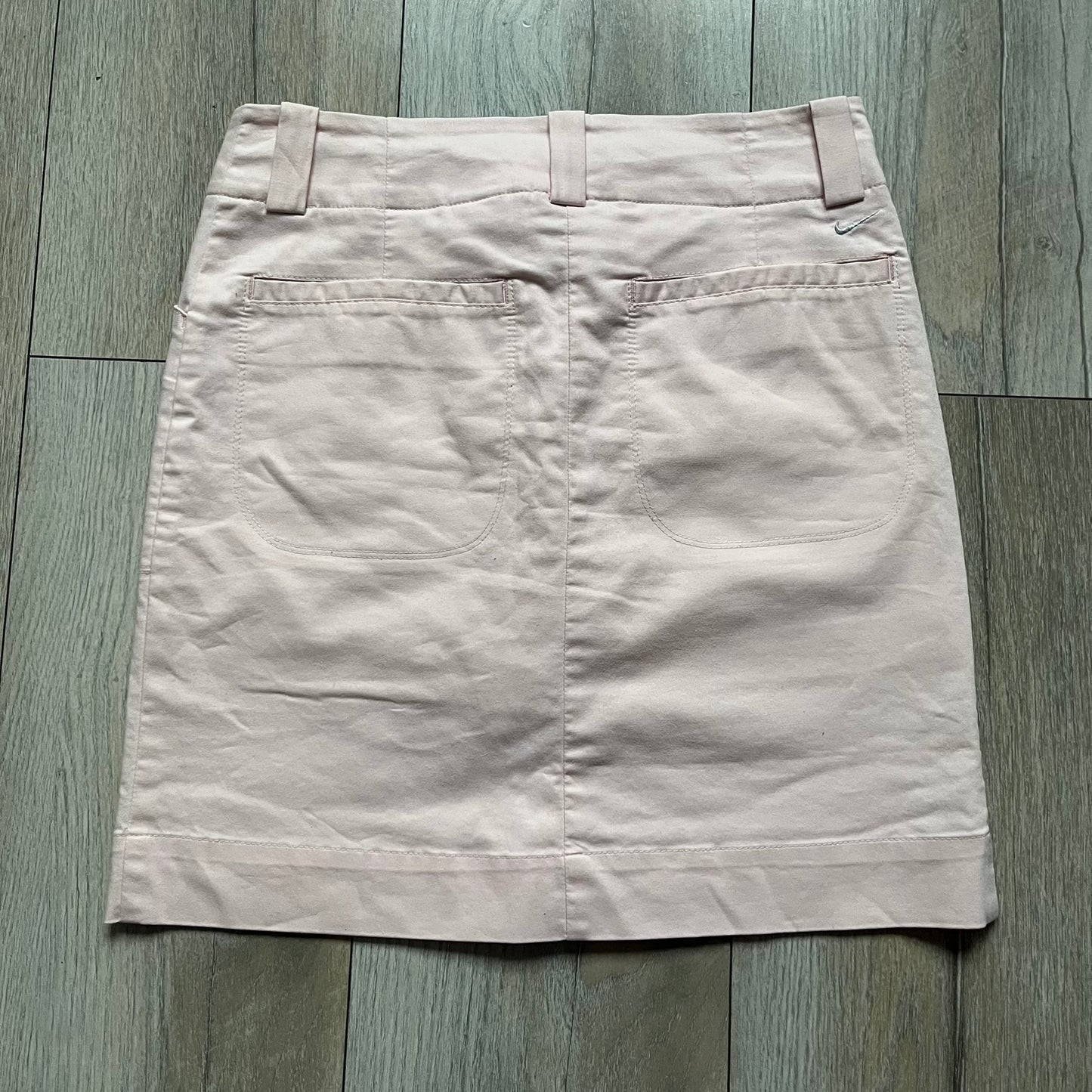 Nike Fit-Dry Light Pink Golf Skirt is a women’s golf skort with integrated inner shorts. The light pink skirt features belt loops at the waistband, front and back pockets, side cutouts for flexibility, and a button closure. A small embroidered Nike logo is placed at the back, maintaining a minimal design.
