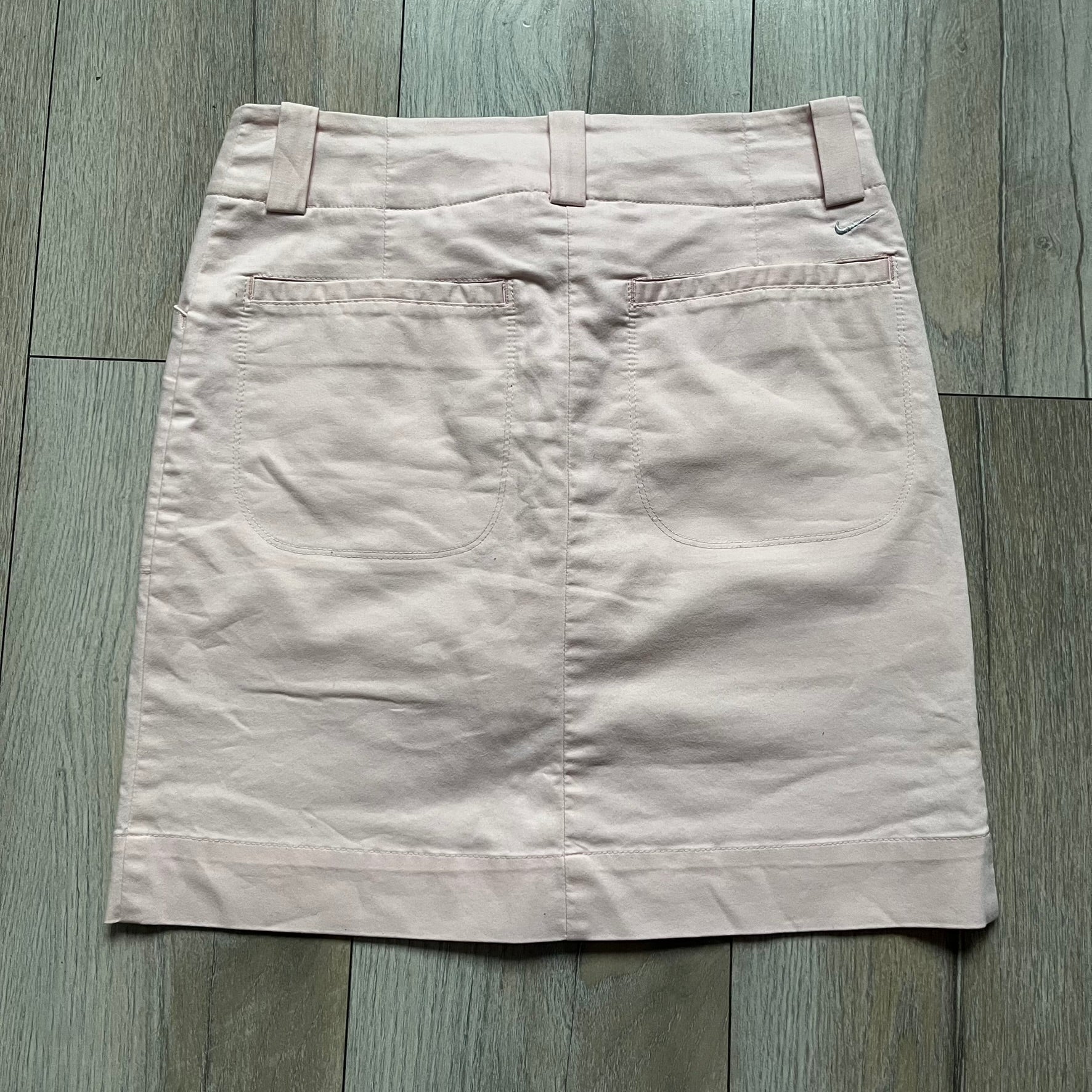 Nike Fit-Dry Light Pink Golf Skirt is a women’s golf skort with integrated inner shorts. The light pink skirt features belt loops at the waistband, front and back pockets, side cutouts for flexibility, and a button closure. A small embroidered Nike logo is placed at the back, maintaining a minimal design.