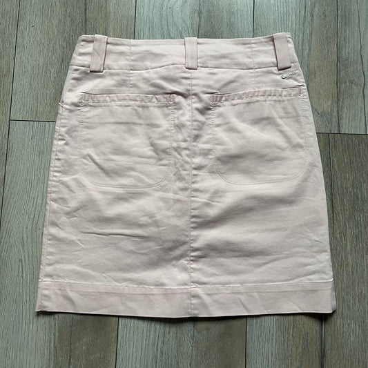 Nike Fit-Dry Light Pink Golf Skirt is a women’s golf skort with integrated inner shorts. The light pink skirt features belt loops at the waistband, front and back pockets, side cutouts for flexibility, and a button closure. A small embroidered Nike logo is placed at the back, maintaining a minimal design.