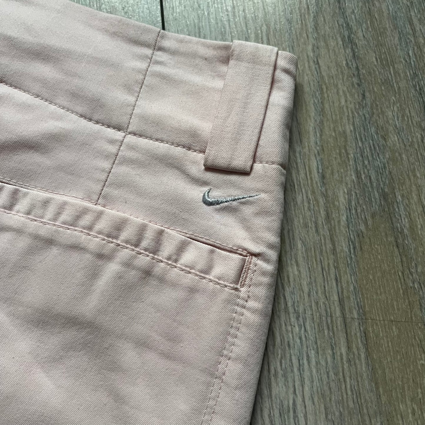 Nike Fit-Dry Light Pink Golf Skirt is a women’s golf skort with integrated inner shorts. The light pink skirt features belt loops at the waistband, front and back pockets, side cutouts for flexibility, and a button closure. A small embroidered Nike logo is placed at the back, maintaining a minimal design.