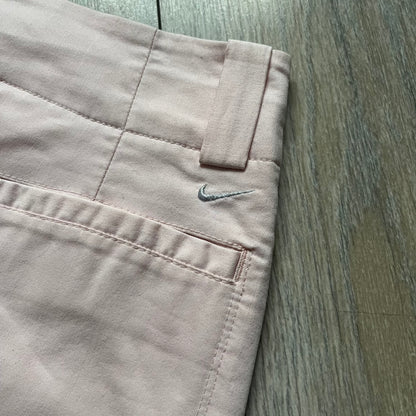 Nike Fit-Dry Light Pink Golf Skirt is a women’s golf skort with integrated inner shorts. The light pink skirt features belt loops at the waistband, front and back pockets, side cutouts for flexibility, and a button closure. A small embroidered Nike logo is placed at the back, maintaining a minimal design.