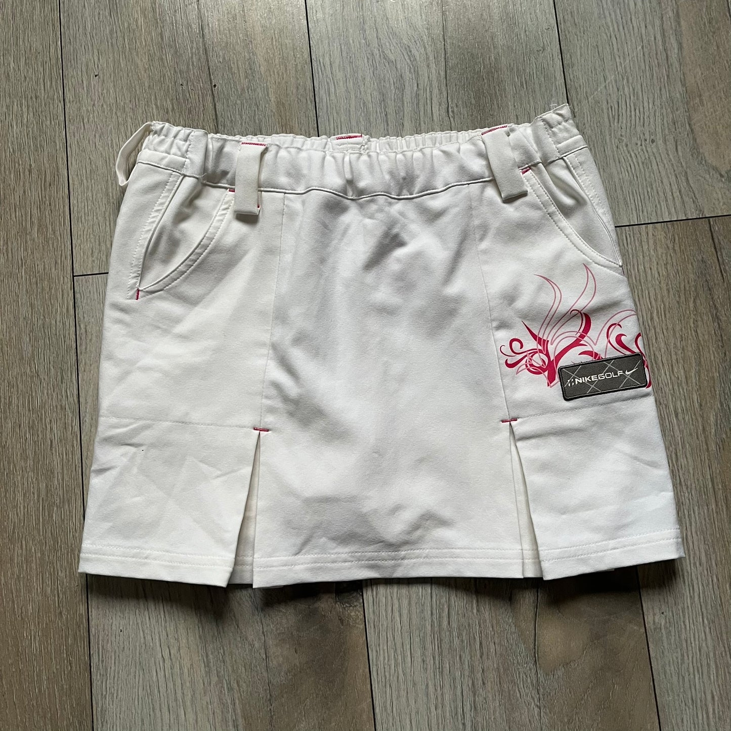 Nike Fit Dry White/Pink Golf Skirt is a women’s golf skort with built-in inner shorts. The white skirt features belt loops at the waistband, front pockets, a small back pocket, and side cutouts on both sides for flexibility. Below the left front pocket there is a small pink floral-style detail along with a Nike Golf patch.