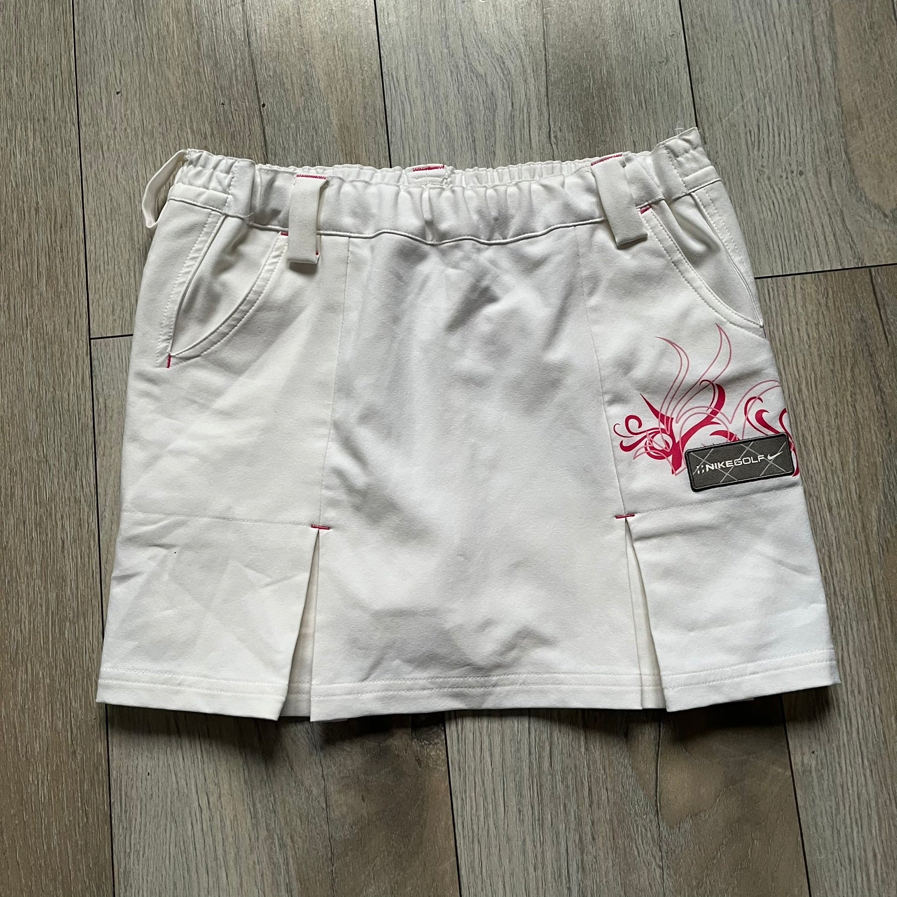 Nike Fit Dry White/Pink Golf Skirt is a women’s golf skort with built-in inner shorts. The white skirt features belt loops at the waistband, front pockets, a small back pocket, and side cutouts on both sides for flexibility. Below the left front pocket there is a small pink floral-style detail along with a Nike Golf patch.