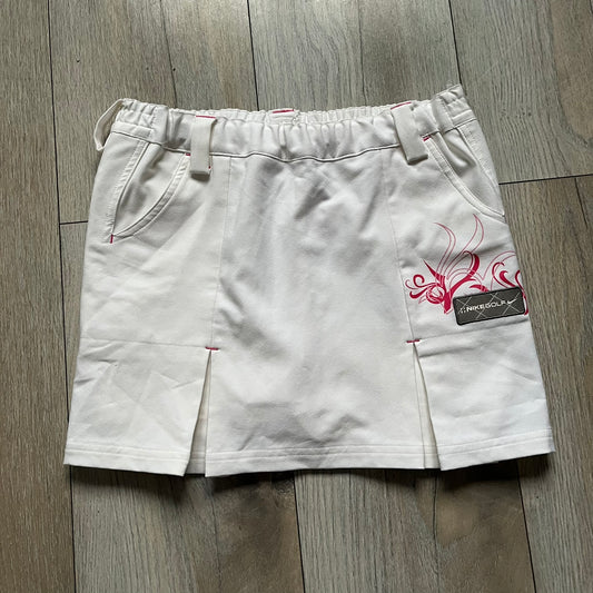 Nike Fit Dry White/Pink Golf Skirt is a women’s golf skort with built-in inner shorts. The white skirt features belt loops at the waistband, front pockets, a small back pocket, and side cutouts on both sides for flexibility. Below the left front pocket there is a small pink floral-style detail along with a Nike Golf patch.