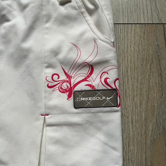 Nike Fit Dry White/Pink Golf Skirt is a women’s golf skort with built-in inner shorts. The white skirt features belt loops at the waistband, front pockets, a small back pocket, and side cutouts on both sides for flexibility. Below the left front pocket there is a small pink floral-style detail along with a Nike Golf patch.