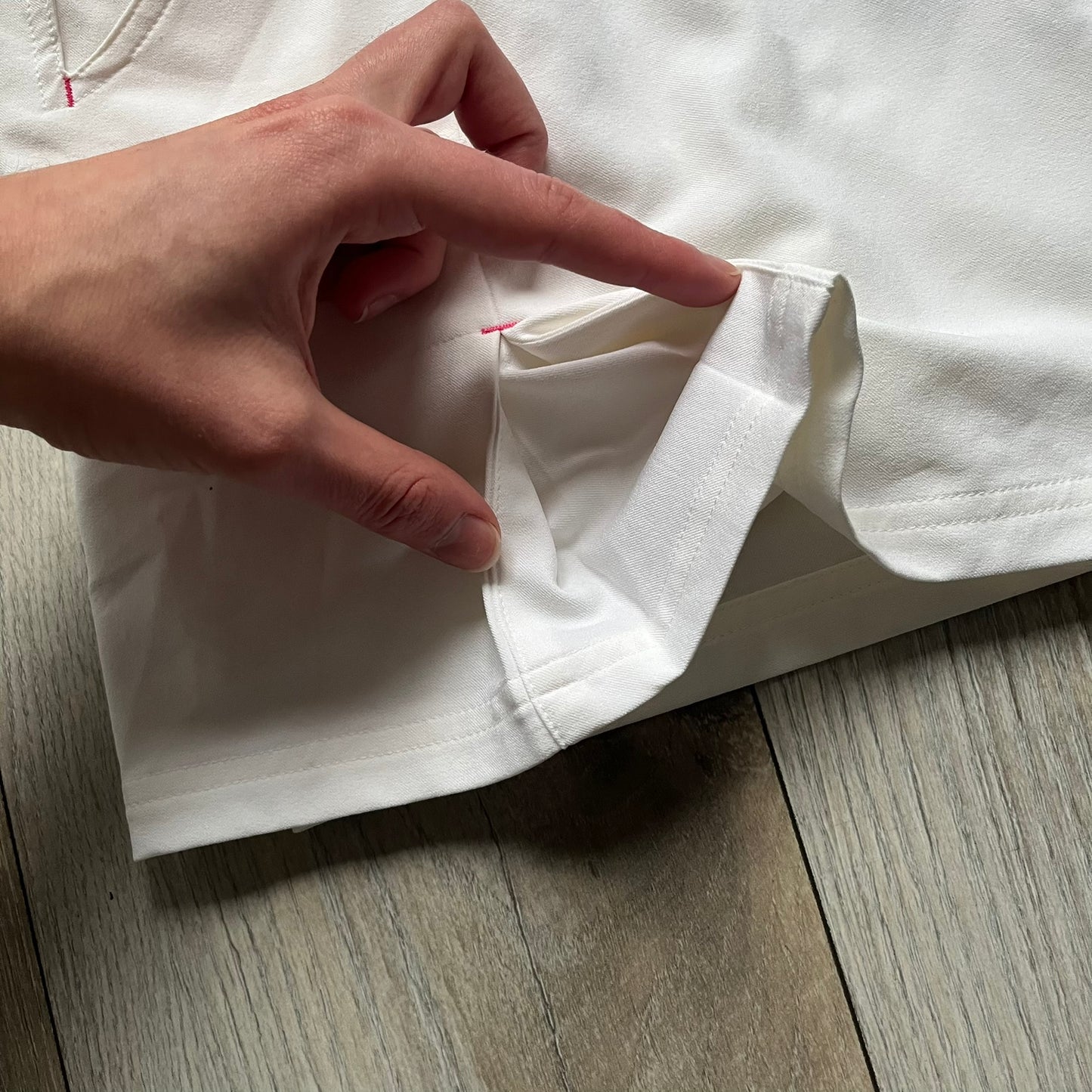 Nike Fit Dry White/Pink Golf Skirt is a women’s golf skort with built-in inner shorts. The white skirt features belt loops at the waistband, front pockets, a small back pocket, and side cutouts on both sides for flexibility. Below the left front pocket there is a small pink floral-style detail along with a Nike Golf patch.