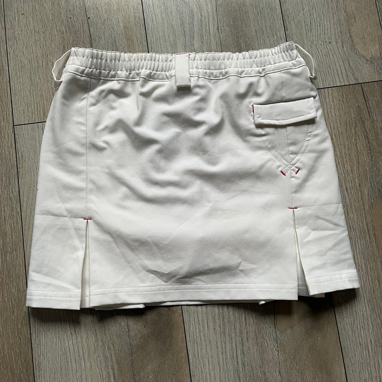 Nike Fit Dry White/Pink Golf Skirt is a women’s golf skort with built-in inner shorts. The white skirt features belt loops at the waistband, front pockets, a small back pocket, and side cutouts on both sides for flexibility. Below the left front pocket there is a small pink floral-style detail along with a Nike Golf patch.