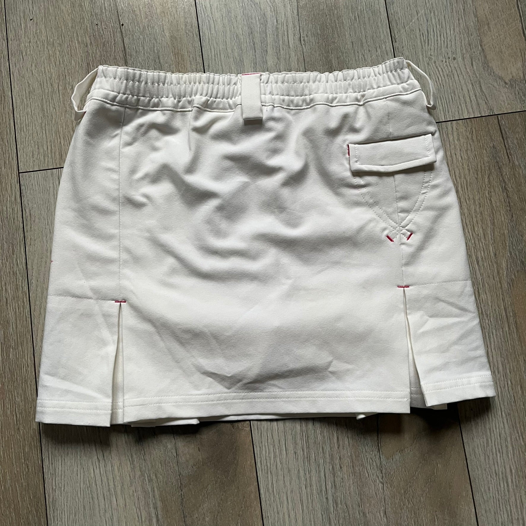 Nike Fit Dry White/Pink Golf Skirt is a women’s golf skort with built-in inner shorts. The white skirt features belt loops at the waistband, front pockets, a small back pocket, and side cutouts on both sides for flexibility. Below the left front pocket there is a small pink floral-style detail along with a Nike Golf patch.