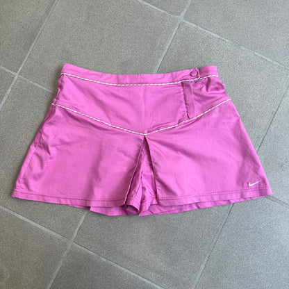 Nike Fuchsia Pleated Skirt features a vibrant fuchsia colorway with a pleated design and a center slit for improved freedom of movement. It is detailed with a small white Nike Swoosh near the hem and a Nike Tennis logo at the waistband. The skirt fastens with a button and zip closure and is finished with contrasting white stitching for a sporty look.