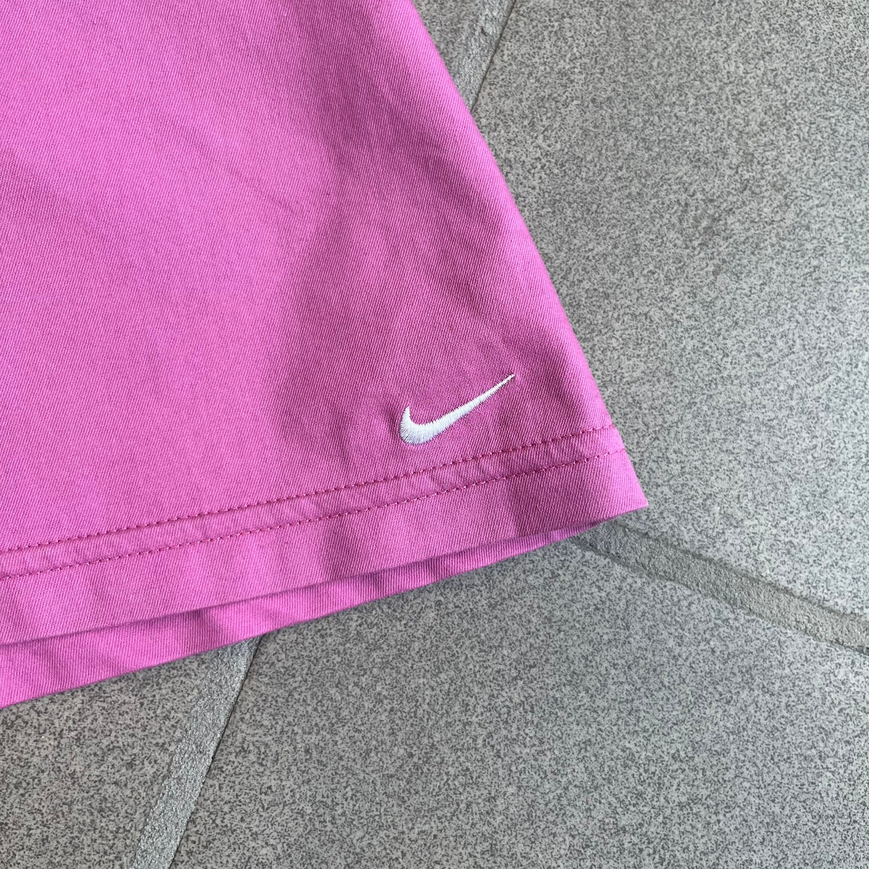 Nike Fuchsia Pleated Skirt features a vibrant fuchsia colorway with a pleated design and a center slit for improved freedom of movement. It is detailed with a small white Nike Swoosh near the hem and a Nike Tennis logo at the waistband. The skirt fastens with a button and zip closure and is finished with contrasting white stitching for a sporty look.