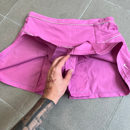 Nike Fuchsia Pleated Skirt features a vibrant fuchsia colorway with a pleated design and a center slit for improved freedom of movement. It is detailed with a small white Nike Swoosh near the hem and a Nike Tennis logo at the waistband. The skirt fastens with a button and zip closure and is finished with contrasting white stitching for a sporty look.