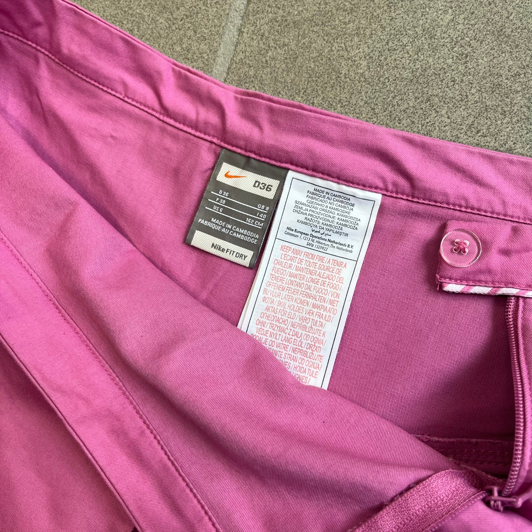 Nike Fuchsia Pleated Skirt features a vibrant fuchsia colorway with a pleated design and a center slit for improved freedom of movement. It is detailed with a small white Nike Swoosh near the hem and a Nike Tennis logo at the waistband. The skirt fastens with a button and zip closure and is finished with contrasting white stitching for a sporty look.
