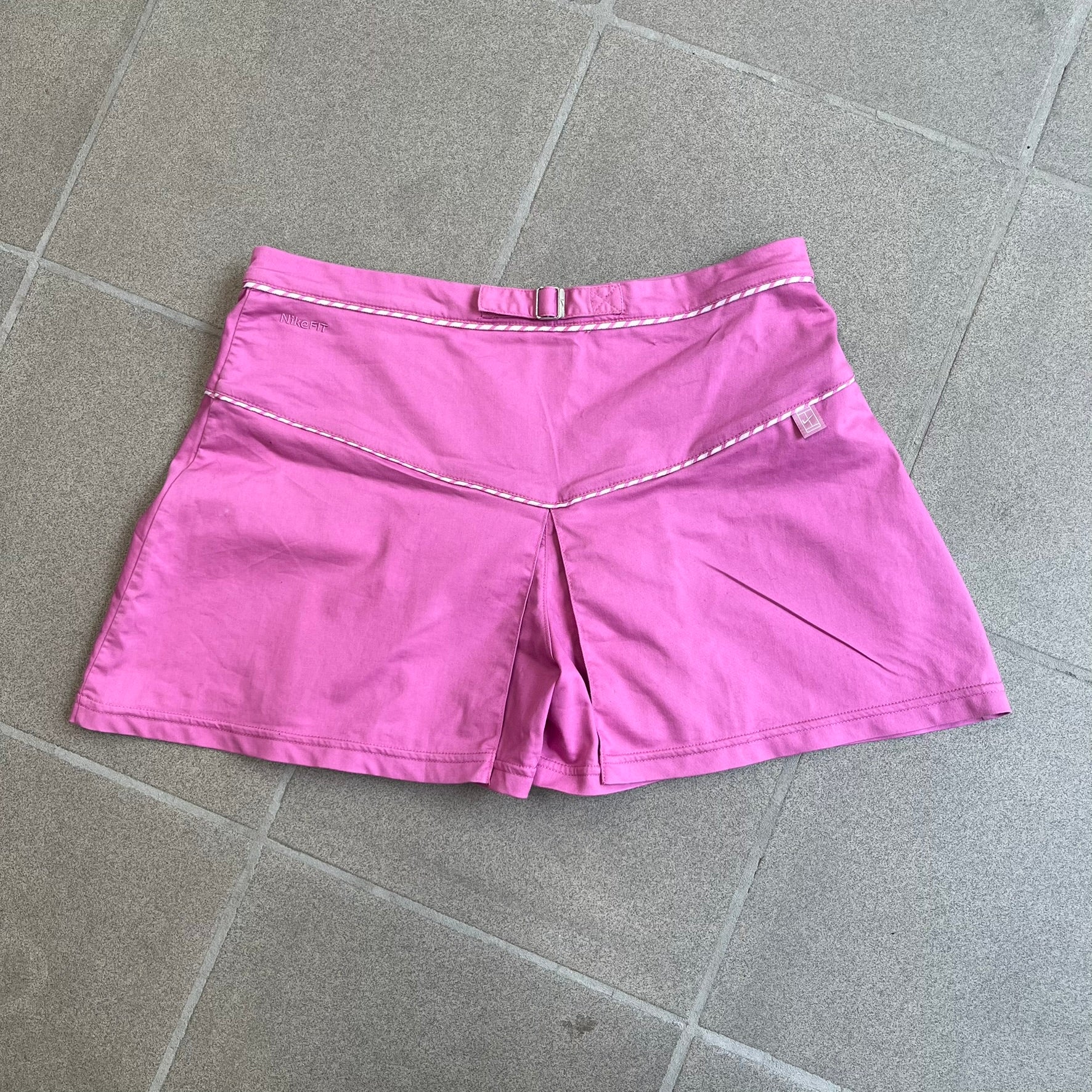 Nike Fuchsia Pleated Skirt features a vibrant fuchsia colorway with a pleated design and a center slit for improved freedom of movement. It is detailed with a small white Nike Swoosh near the hem and a Nike Tennis logo at the waistband. The skirt fastens with a button and zip closure and is finished with contrasting white stitching for a sporty look.