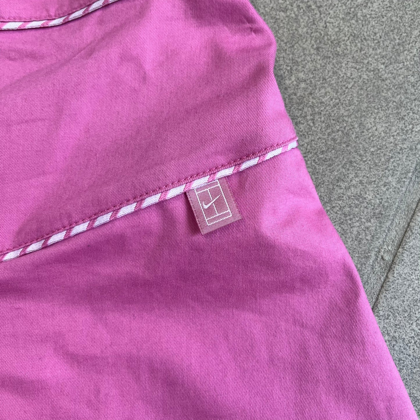 Nike Fuchsia Pleated Skirt features a vibrant fuchsia colorway with a pleated design and a center slit for improved freedom of movement. It is detailed with a small white Nike Swoosh near the hem and a Nike Tennis logo at the waistband. The skirt fastens with a button and zip closure and is finished with contrasting white stitching for a sporty look.