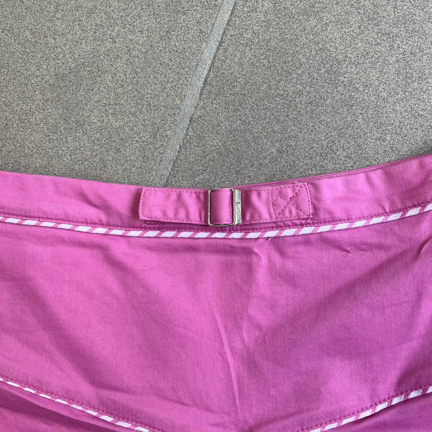 Nike Fuchsia Pleated Skirt features a vibrant fuchsia colorway with a pleated design and a center slit for improved freedom of movement. It is detailed with a small white Nike Swoosh near the hem and a Nike Tennis logo at the waistband. The skirt fastens with a button and zip closure and is finished with contrasting white stitching for a sporty look.