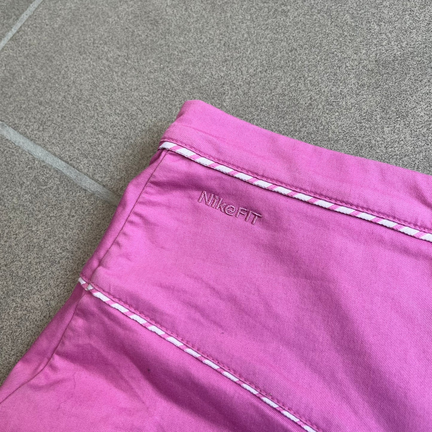 Nike Fuchsia Pleated Skirt features a vibrant fuchsia colorway with a pleated design and a center slit for improved freedom of movement. It is detailed with a small white Nike Swoosh near the hem and a Nike Tennis logo at the waistband. The skirt fastens with a button and zip closure and is finished with contrasting white stitching for a sporty look.