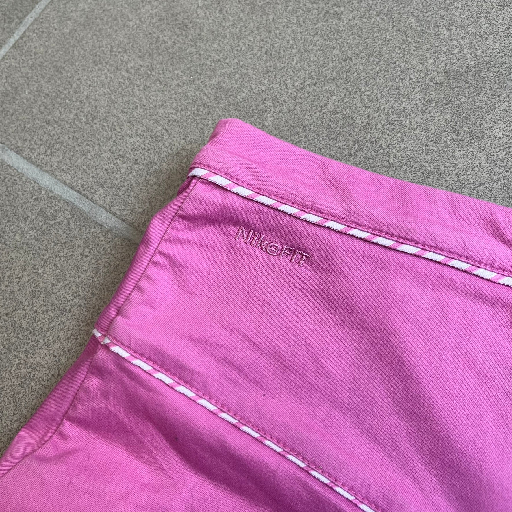 Nike Fuchsia Pleated Skirt features a vibrant fuchsia colorway with a pleated design and a center slit for improved freedom of movement. It is detailed with a small white Nike Swoosh near the hem and a Nike Tennis logo at the waistband. The skirt fastens with a button and zip closure and is finished with contrasting white stitching for a sporty look.