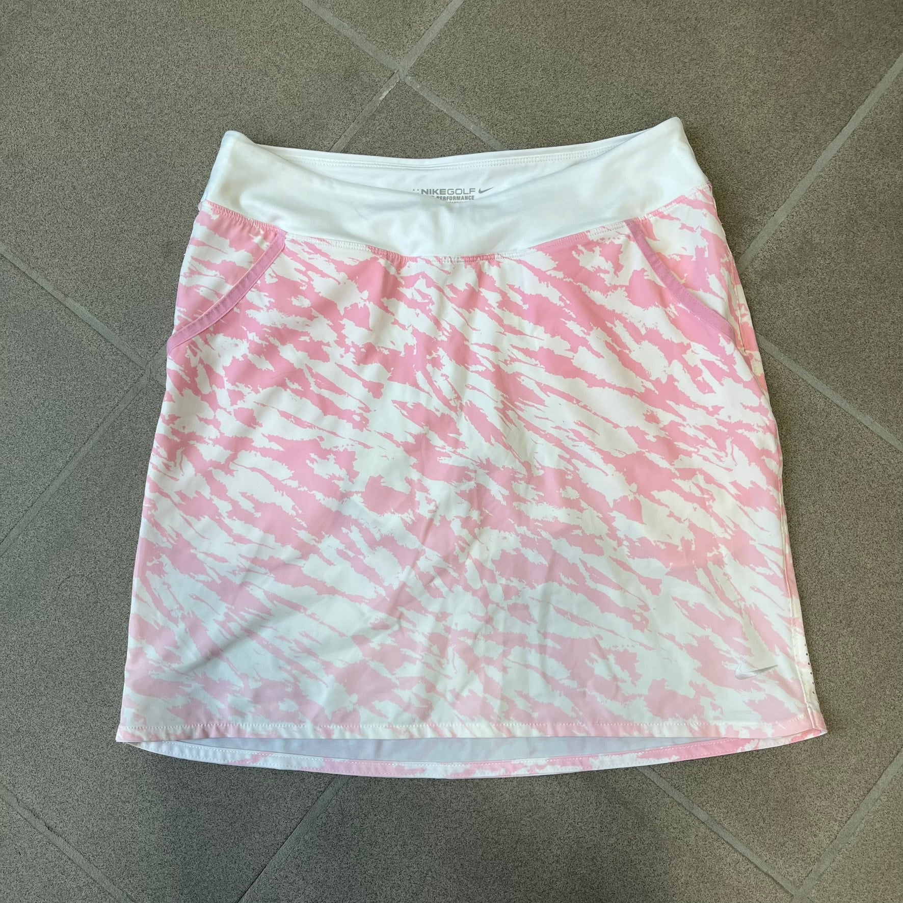 Nike Golf Pink & White Animal Print Skort features a white base with a pink animal-style camouflage pattern that gradually fades toward the lower section. It has built-in white shorts underneath, an embroidered Nike logo at the bottom corner, and pockets on both the front and back.
