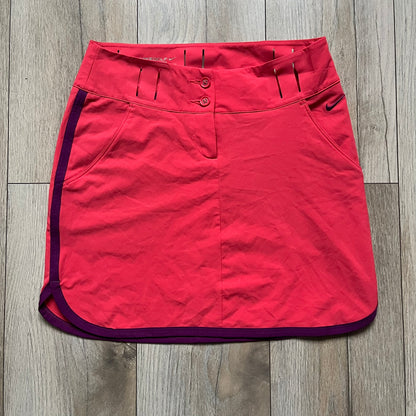 Nike Golf Tour Performance Golf Skirt is a women’s golf skirt in dark pink with belt loops integrated into the waistband. The side and lower hem outlines are finished in purple, and the skirt features front and back pockets with a button closure. An embroidered Nike logo is placed at the front
