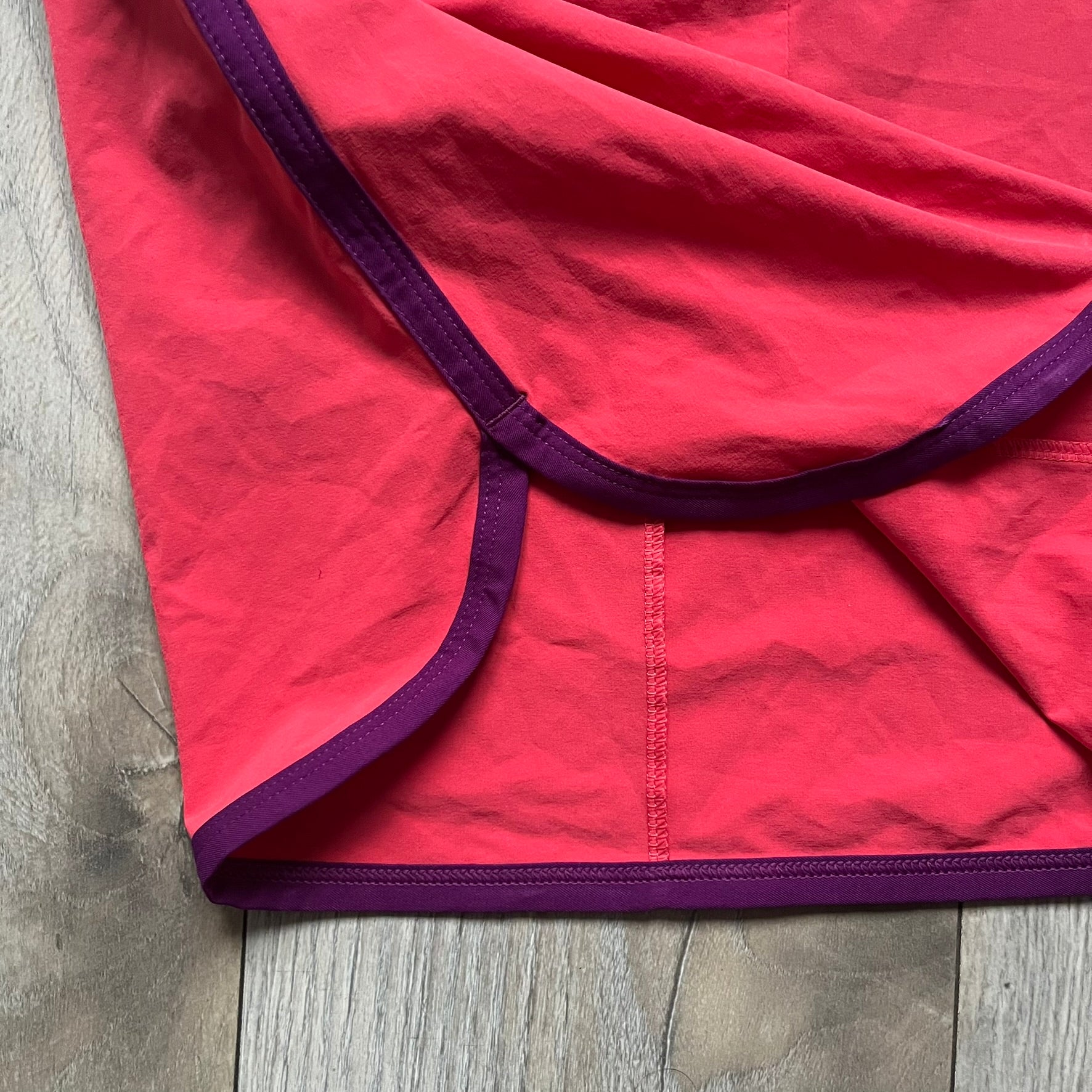 Nike Golf Tour Performance Golf Skirt is a women’s golf skirt in dark pink with belt loops integrated into the waistband. The side and lower hem outlines are finished in purple, and the skirt features front and back pockets with a button closure. An embroidered Nike logo is placed at the front