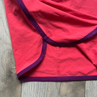 Nike Golf Tour Performance Golf Skirt is a women’s golf skirt in dark pink with belt loops integrated into the waistband. The side and lower hem outlines are finished in purple, and the skirt features front and back pockets with a button closure. An embroidered Nike logo is placed at the front