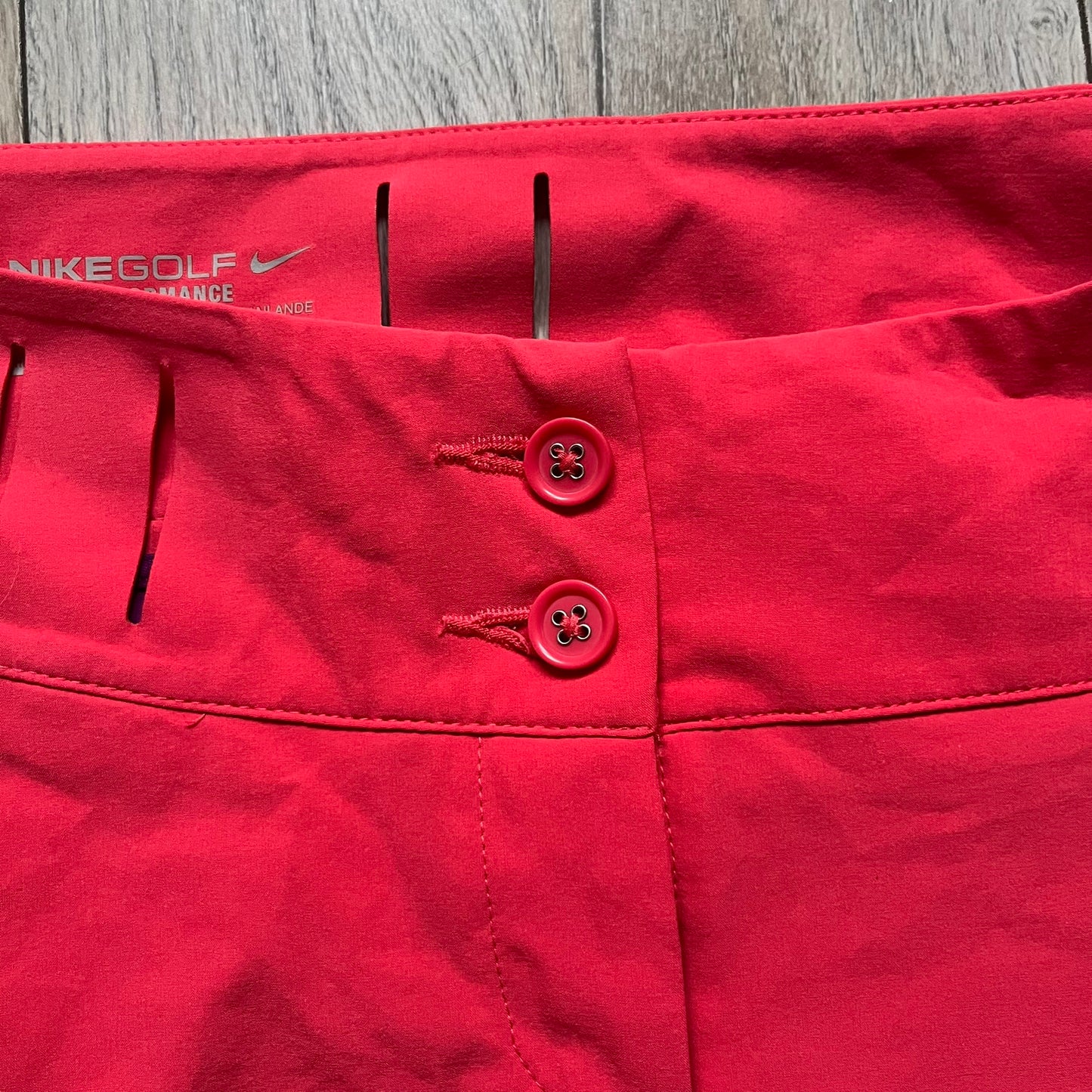 Nike Golf Tour Performance Golf Skirt is a women’s golf skirt in dark pink with belt loops integrated into the waistband. The side and lower hem outlines are finished in purple, and the skirt features front and back pockets with a button closure. An embroidered Nike logo is placed at the front