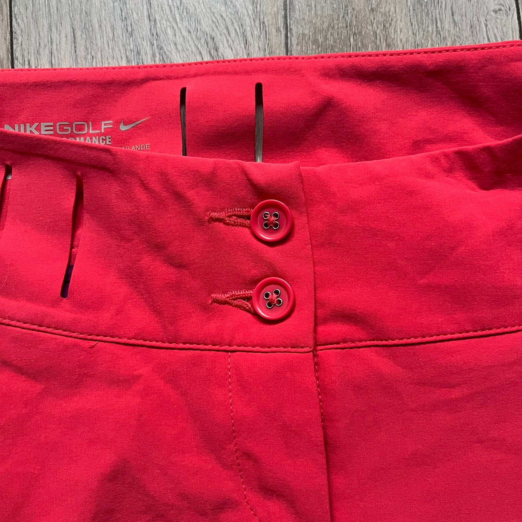 Nike Golf Tour Performance Golf Skirt is a women’s golf skirt in dark pink with belt loops integrated into the waistband. The side and lower hem outlines are finished in purple, and the skirt features front and back pockets with a button closure. An embroidered Nike logo is placed at the front