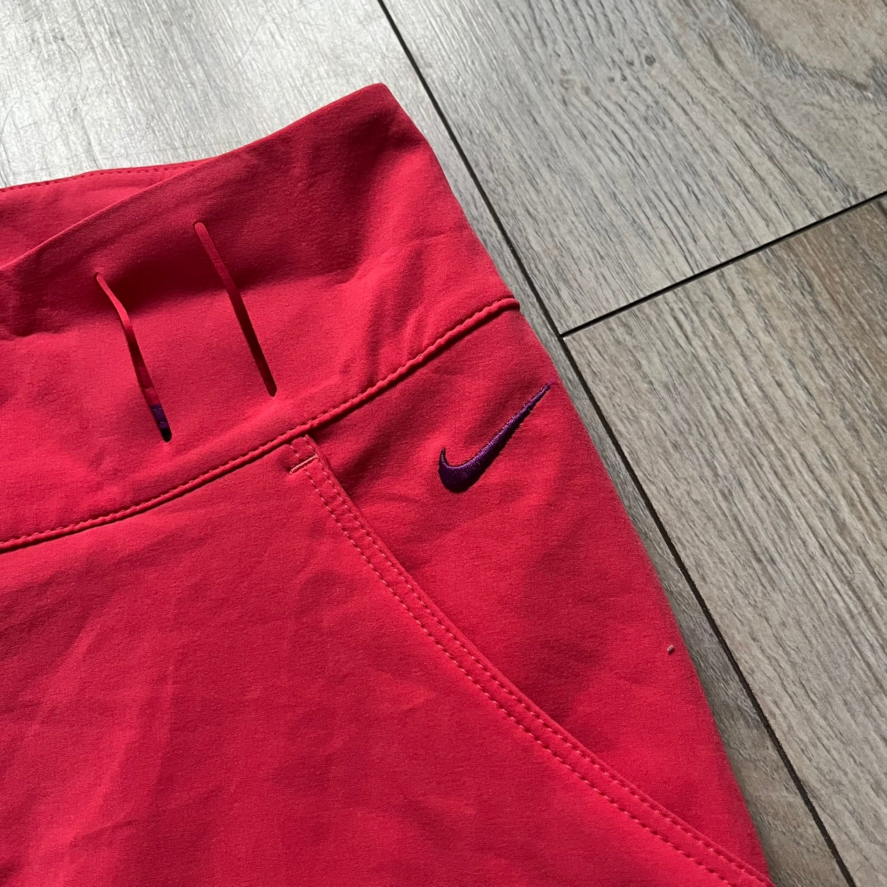 Nike Golf Tour Performance Golf Skirt is a women’s golf skirt in dark pink with belt loops integrated into the waistband. The side and lower hem outlines are finished in purple, and the skirt features front and back pockets with a button closure. An embroidered Nike logo is placed at the front