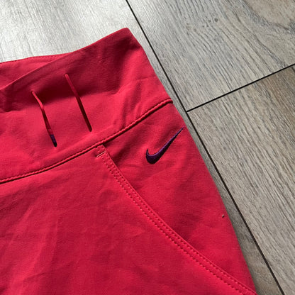 Nike Golf Tour Performance Golf Skirt is a women’s golf skirt in dark pink with belt loops integrated into the waistband. The side and lower hem outlines are finished in purple, and the skirt features front and back pockets with a button closure. An embroidered Nike logo is placed at the front
