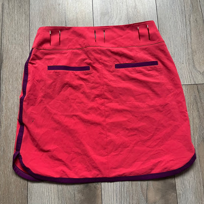 Nike Golf Tour Performance Golf Skirt is a women’s golf skirt in dark pink with belt loops integrated into the waistband. The side and lower hem outlines are finished in purple, and the skirt features front and back pockets with a button closure. An embroidered Nike logo is placed at the front