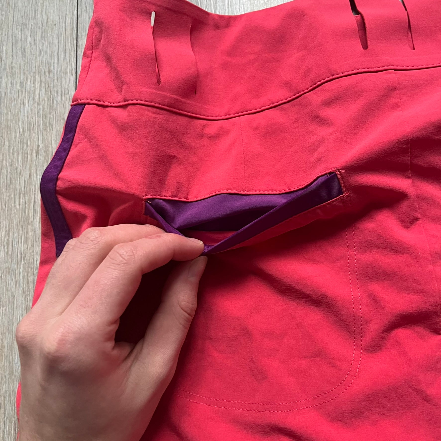 Nike Golf Tour Performance Golf Skirt is a women’s golf skirt in dark pink with belt loops integrated into the waistband. The side and lower hem outlines are finished in purple, and the skirt features front and back pockets with a button closure. An embroidered Nike logo is placed at the front