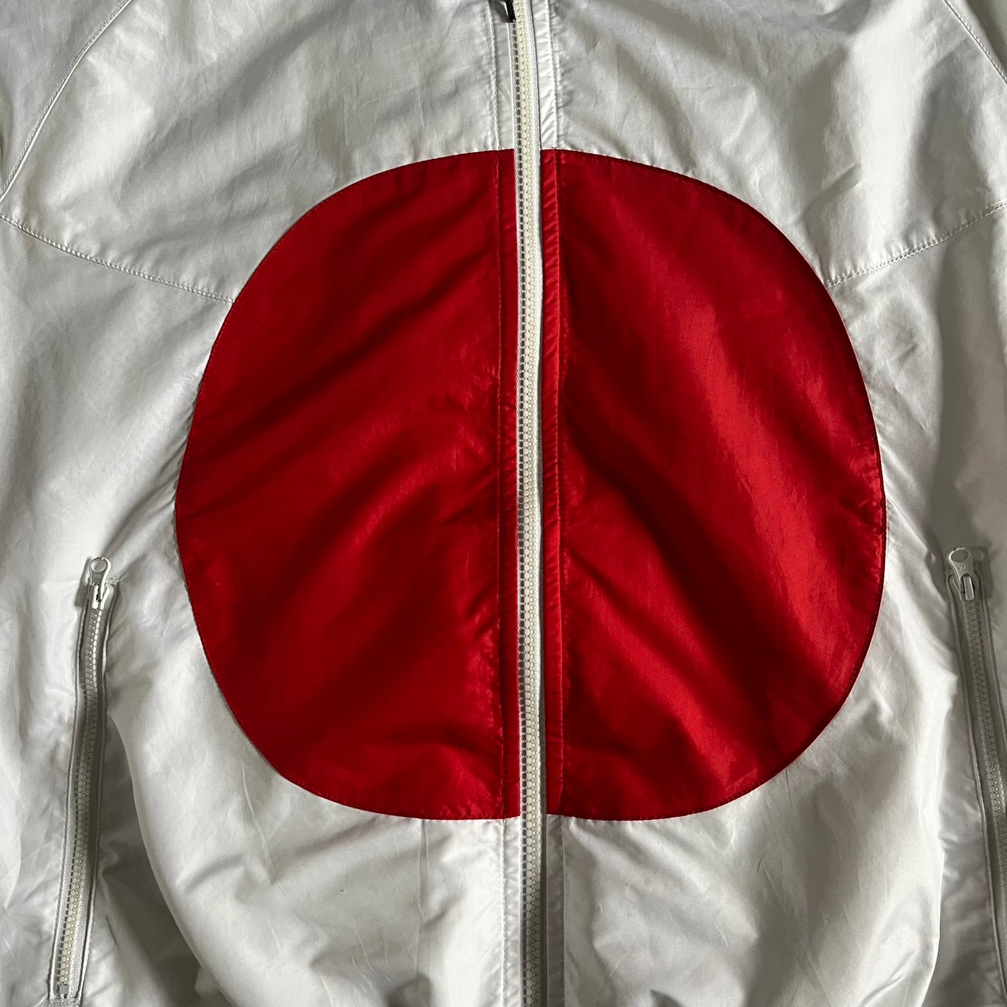 Nike Japan Olympics Split Windbreaker from 2008, created by designer Jarrett Reynolds. Crafted in white with a large red circle representing Japan’s flag, this innovative jacket can be zipped apart on both sides and merged with other compatible models. Hidden inside, a small Japan flag accent completes the concept.