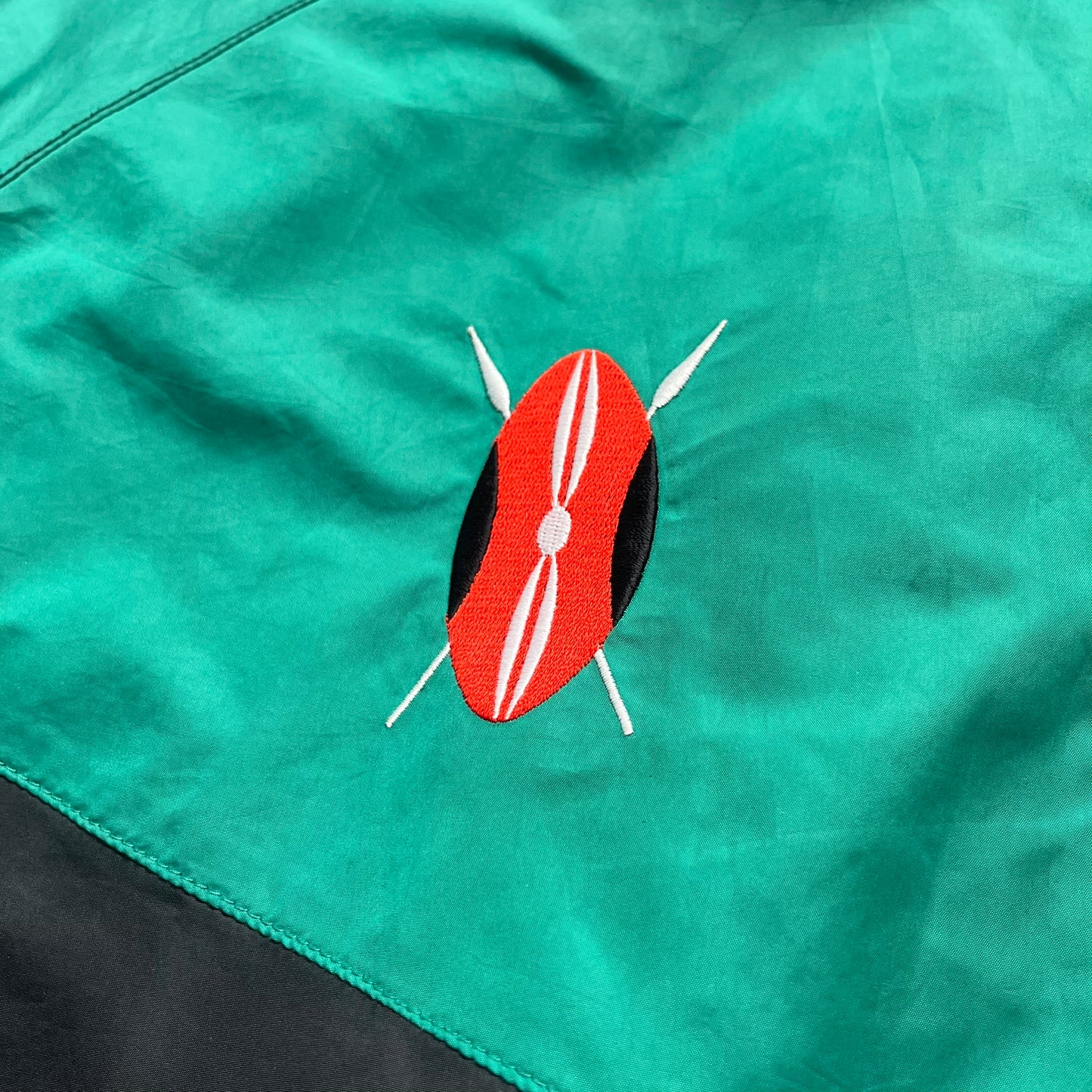 Nike Kenya Olympics Split Windbreaker from 2008, created by designer Jarrett Reynolds. Crafted in black and green. Small Maasai shield on one side of the chest with Nike red embroidery on the other. The upper half of the jacket is green, bottom and arms are fully black. This innovative jacket can be zipped apart on both sides and merged with other compatible models. Hidden inside, a small Kenya flags accent completes the concept.