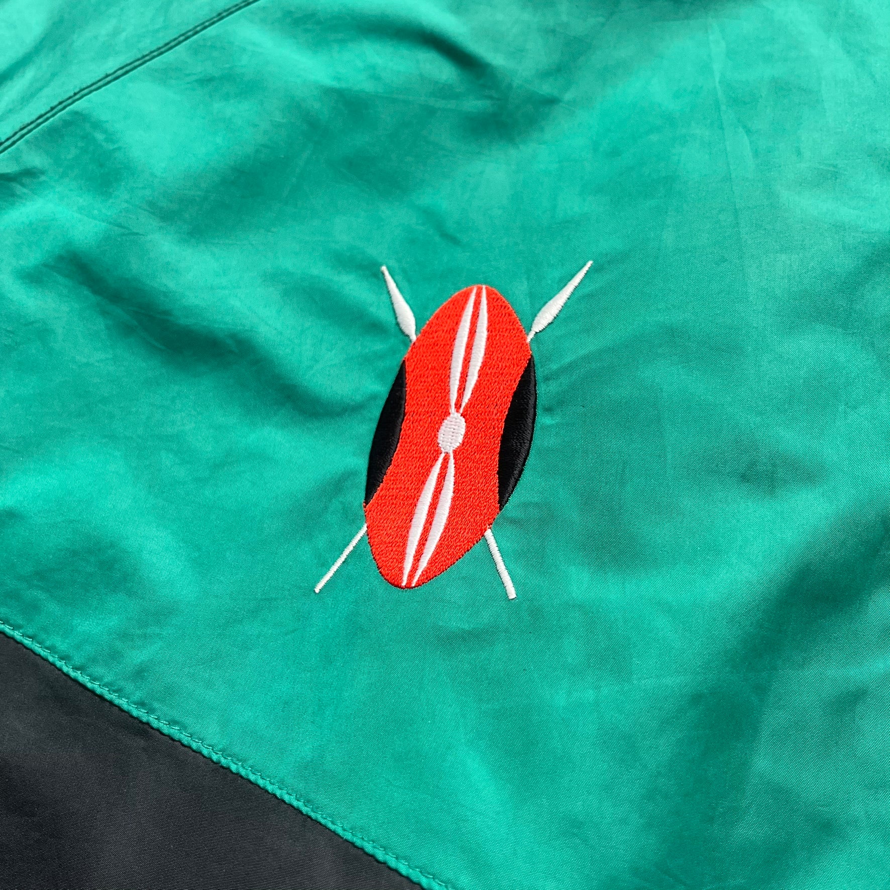 Nike Kenya Olympics Split Windbreaker from 2008, created by designer Jarrett Reynolds. Crafted in black and green. Small Maasai shield on one side of the chest with Nike red embroidery on the other. The upper half of the jacket is green, bottom and arms are fully black. This innovative jacket can be zipped apart on both sides and merged with other compatible models. Hidden inside, a small Kenya flags accent completes the concept.