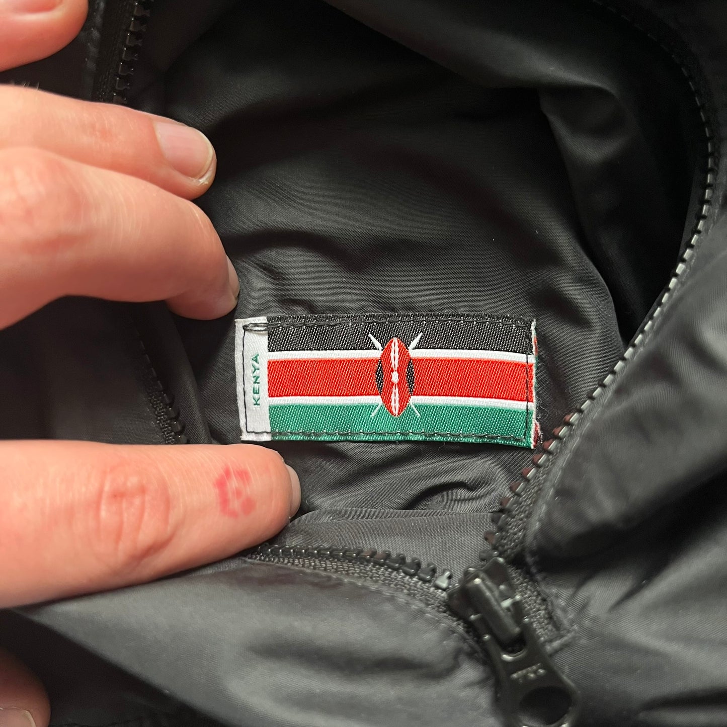 Nike Kenya Olympics Split Windbreaker from 2008, created by designer Jarrett Reynolds. Crafted in black and green. Small Maasai shield on one side of the chest with Nike red embroidery on the other. The upper half of the jacket is green, bottom and arms are fully black. This innovative jacket can be zipped apart on both sides and merged with other compatible models. Hidden inside, a small Kenya flags accent completes the concept.