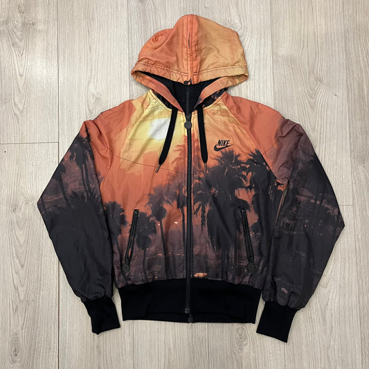 Nike “Beach Sunset” Split Windbreaker WMNS features dual zippers along the front and back, allowing the jacket to separate and merge with other compatible models from the collection. The all-over print shows an orange and yellow sunset with black palms, complemented by black hems and interior lining. Finished with circular metal Nike zip pulls and a small black embroidered Swoosh on the right chest.