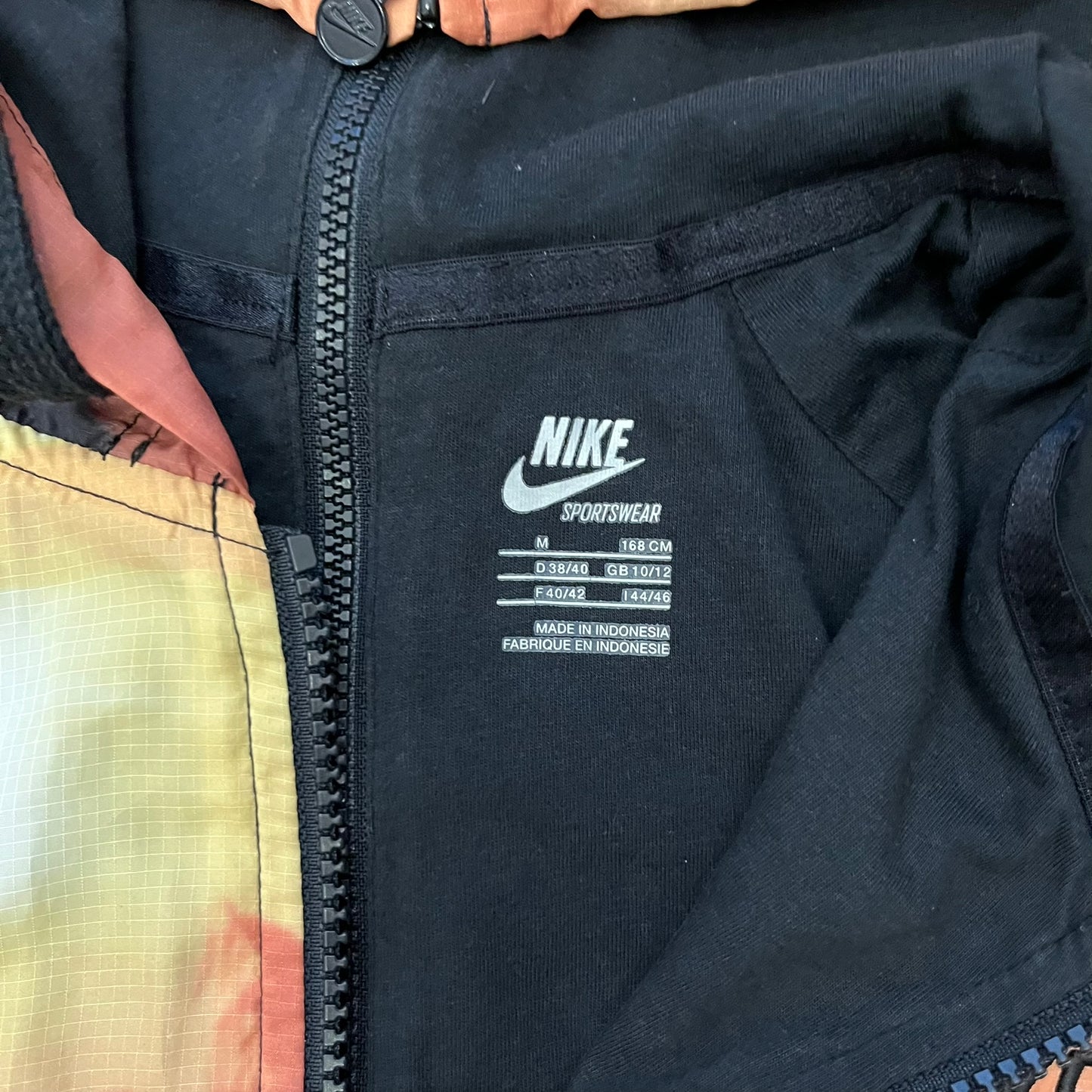 Nike “Beach Sunset” Split Windbreaker WMNS features dual zippers along the front and back, allowing the jacket to separate and merge with other compatible models from the collection. The all-over print shows an orange and yellow sunset with black palms, complemented by black hems and interior lining. Finished with circular metal Nike zip pulls and a small black embroidered Swoosh on the right chest.